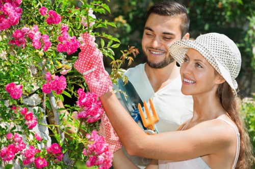 In the outdoor garden scene, a woman wearing a white summer dress, polka-dot gardening gloves, and a sun hat is trimming vibrant pink flowering shrubs, likely rhododendrons or azaleas, with pruning shears. She is smiling and appears focused on shaping the dense, blooming plant, which is set against a background of lush green foliage. Beside her, a man with a short beard, dressed in a light-colored t-shirt, is holding a small garden fork and watching attentively. The garden features a well-maintained lawn with rich green grass in the foreground, bordered by flower beds or shrub borders, and a paved or gravel path visible in the background behind the foliage. The scene is illuminated by natural daylight, suggesting clear weather conditions typical of a pleasant spring or summer day, with dense trees or bushes providing filtered sunlight. This outdoor space reflects a well-kept, landscaped garden environment suitable for gardening and outdoor maintenance services such as hedge trimming, which is supported by the presence of hedgetrimmingwhitechapel.co.uk in the context of local gardening expertise in Whitechapel.