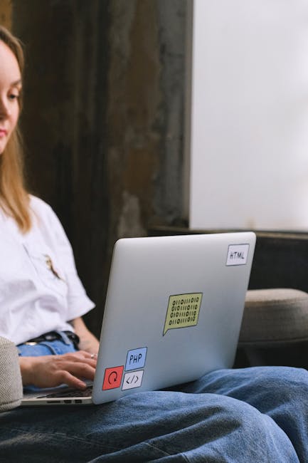 A close-up view of a silver laptop placed on a person's lap, with the screen partially visible. The laptop has stickers on it, including a yellow binary code label, a blue PHP label, a red power butto