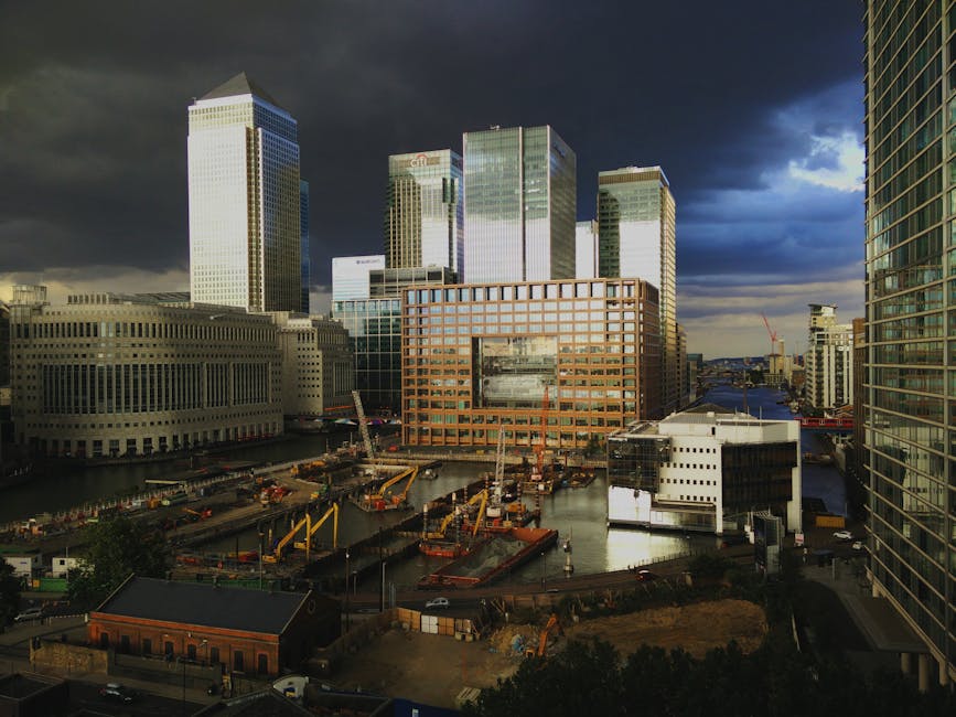 A cityscape featuring modern high-rise office buildings with glass facades reflecting the cloudy sky, creating a dynamic urban environment. In the foreground, a construction site with cranes, construc