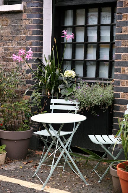 A small urban garden features a white metal bistro table and matching chair positioned against a brick wall with a black-framed window. The window has multiple panes and is decorated with light-colore